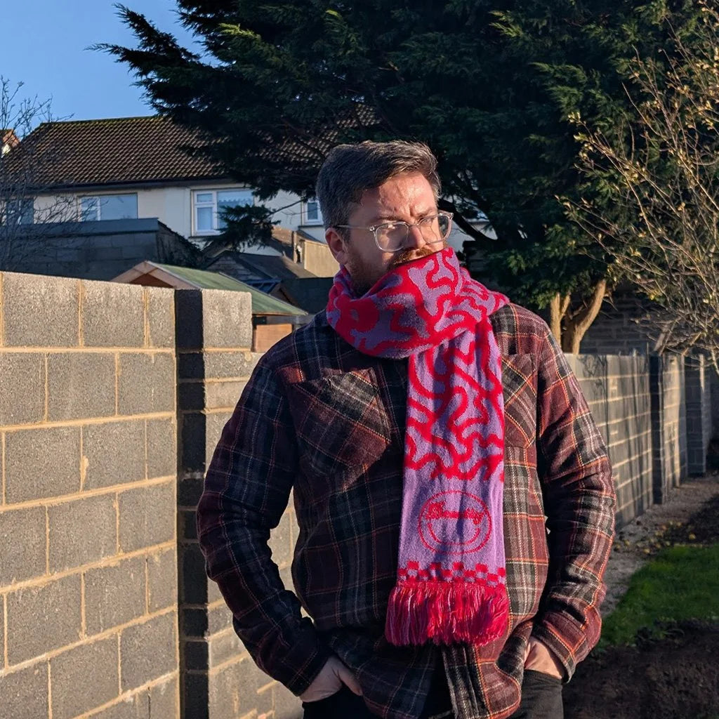 Seán wearing a red and purple patterned scarf in an outdoor setting with a brick wall and trees.