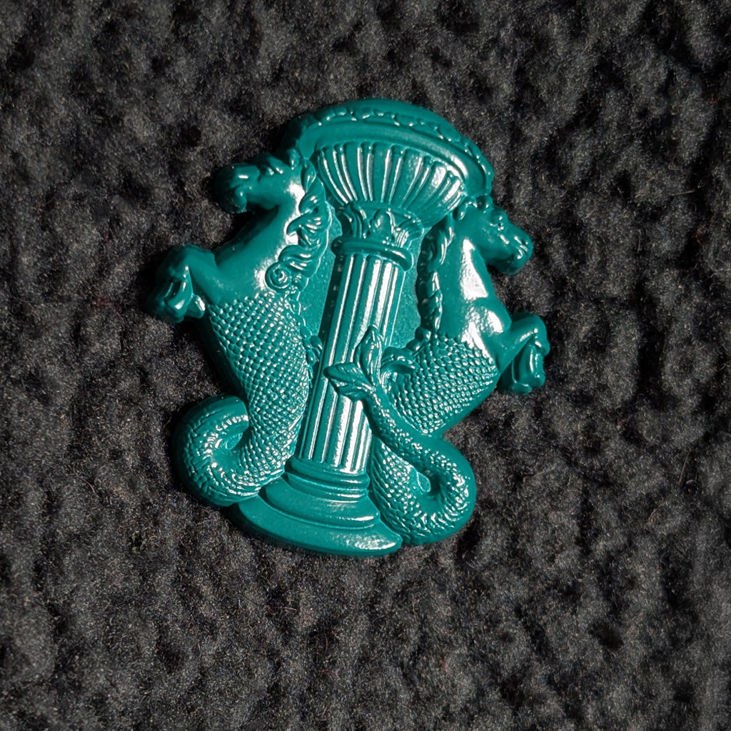 A glossy teal green enamel pin featuring a classical column flanked by two hippocampus, photographed on a dark textured fabric background.