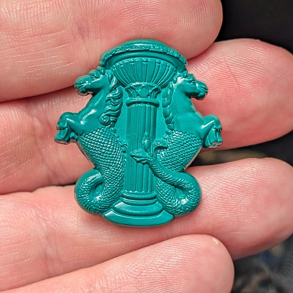 A turquoise green enamel pin featuring two hippocampus wrapping around a central column, held between human fingers.
