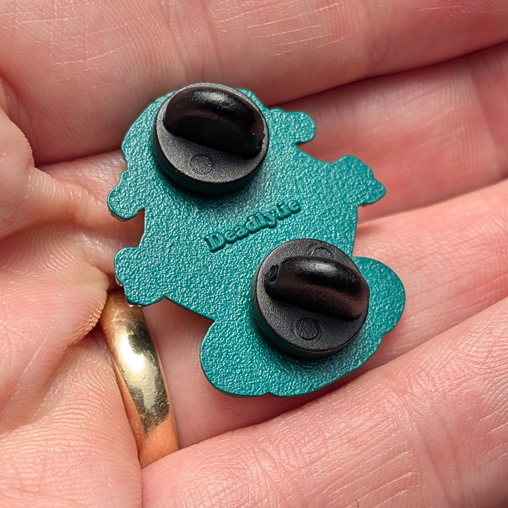 A close-up of a teal and black enamel pin held in a person's hand. The pin features a teal outline shaped like a small creature or blob, with two black oval enamel inserts and the brand name 'Deadly.ie' embossed in the centre.