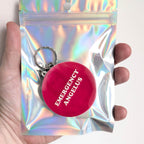 A hand holding a silver holographic resealable pouch containing a round red keychain with the words 'EMERGENCY ANGELUS' in white text.