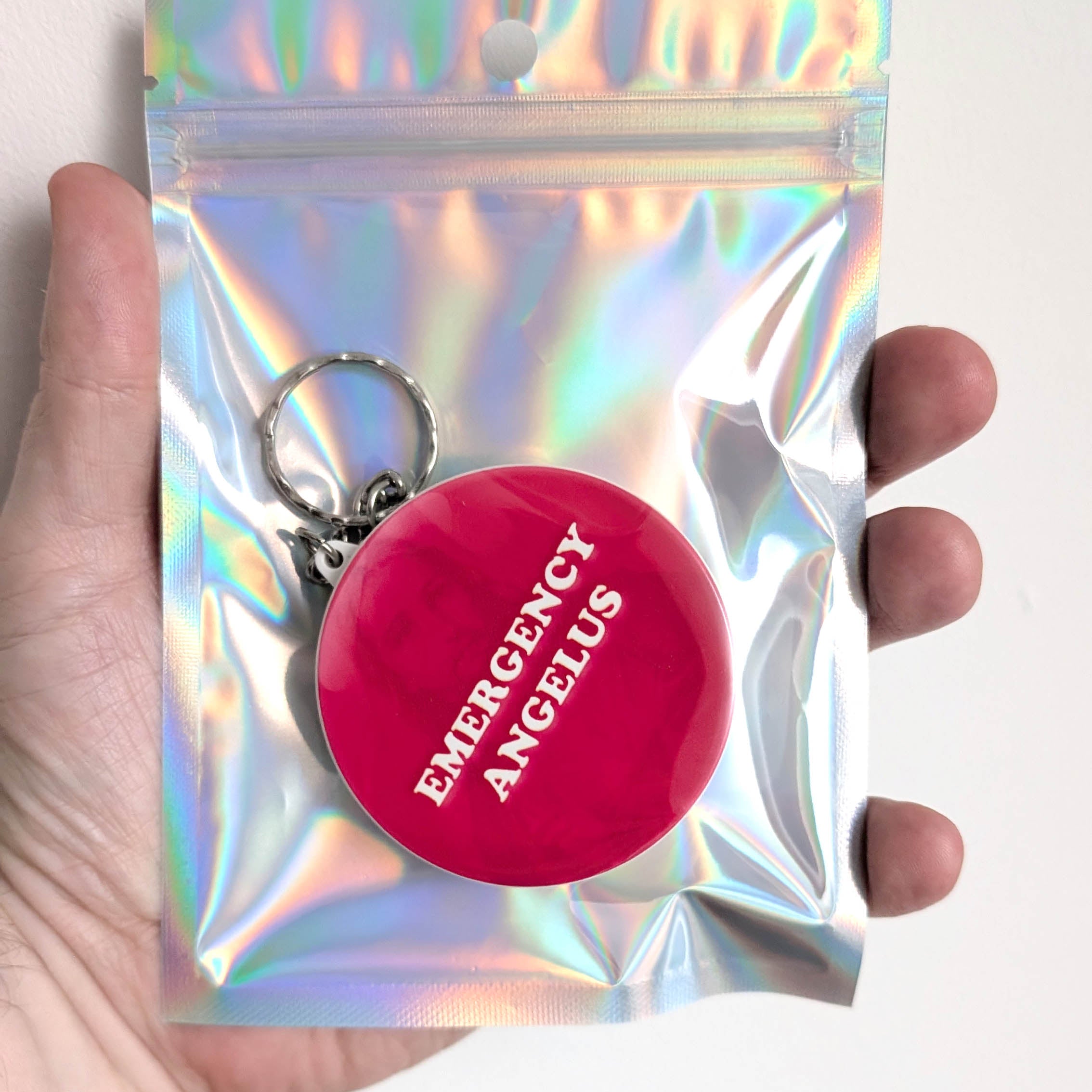 A hand holding a silver holographic resealable pouch containing a round red keychain with the words 'EMERGENCY ANGELUS' in white text.