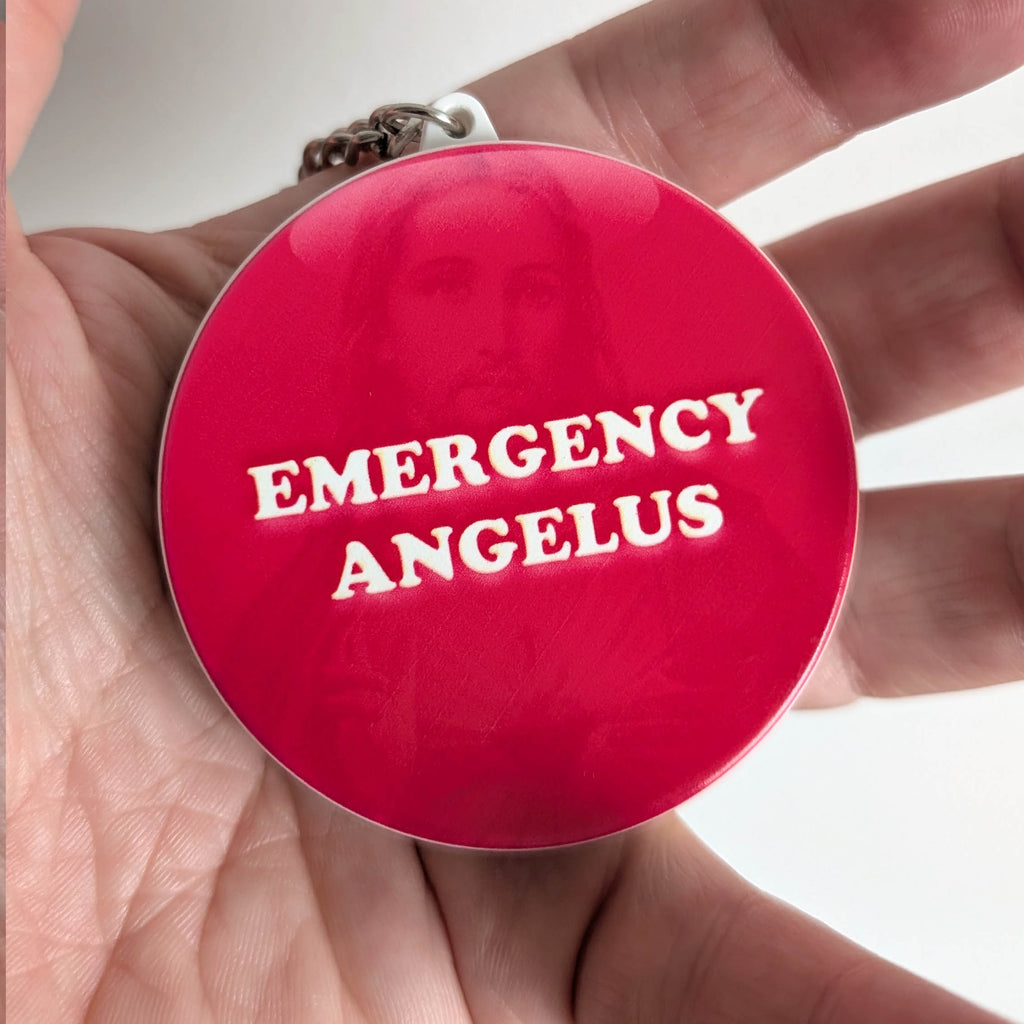 A round red pendant or keychain with the words 'EMERGENCY ANGELUS' in raised white lettering, held in a person's hand. A faint portrait of Jesus is visible through the glossy red surface, and a small metal chain is attached at the top.