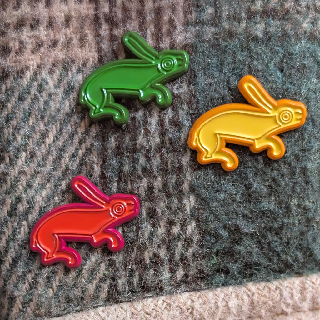 Three Hare Pins, each a different colour of green, yellow and red on a plain green jacket