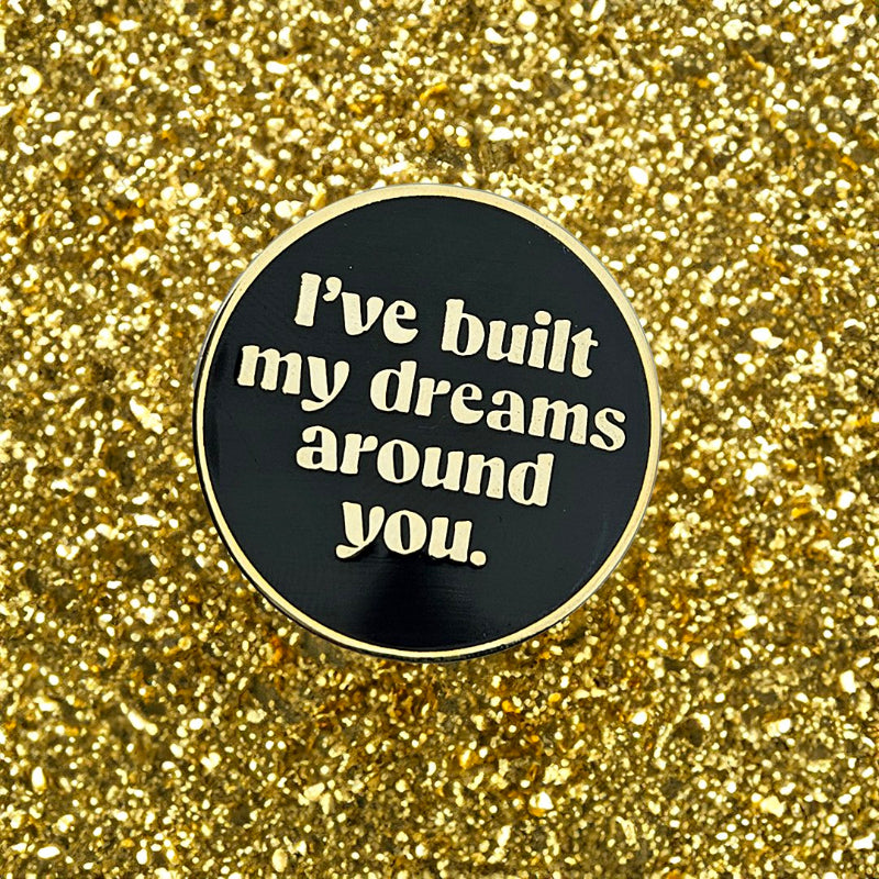 Round black and gold enamel pin with 'I've built my dreams around you' text on a glittery gold background