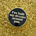 Round black and gold enamel pin with 'I've built my dreams around you' text on a glittery gold background