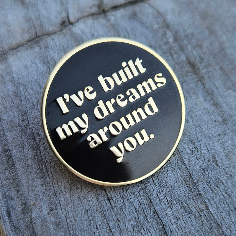 Round black and gold pin with 'I've built my dreams around you' text on a wooden surface