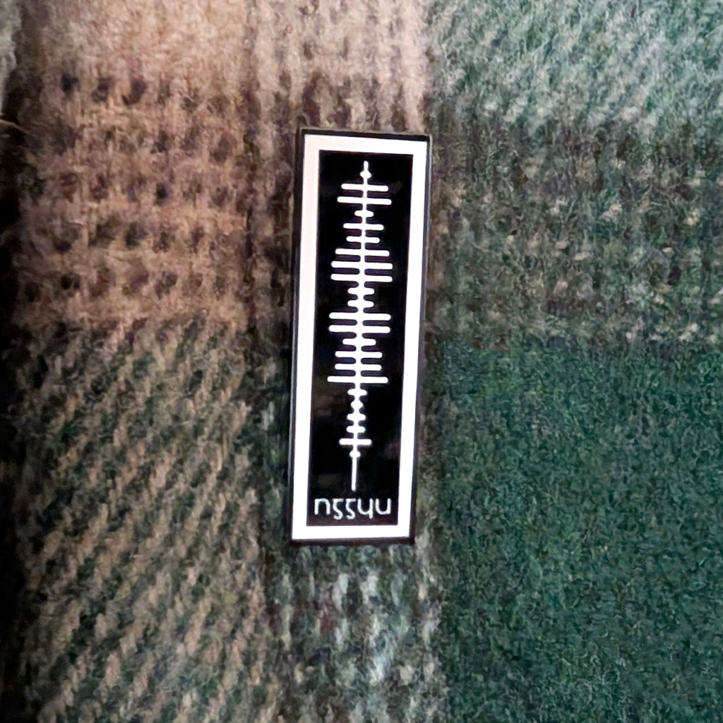 A rectangular black enamel pin with an Ogham graphic and the letters 'NGGYU' at the bottom, displayed on a textured green and brown plaid fabric background.