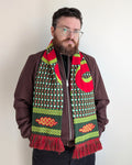 A man with a beard and glasses wearing a brown jacket and black pants, modeling a vibrant patterned knit scarf with red, green, and yellow colors.