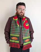 A man with a beard and glasses wearing a brown jacket and black pants, modeling a vibrant patterned knit scarf with red, green, and yellow colors.