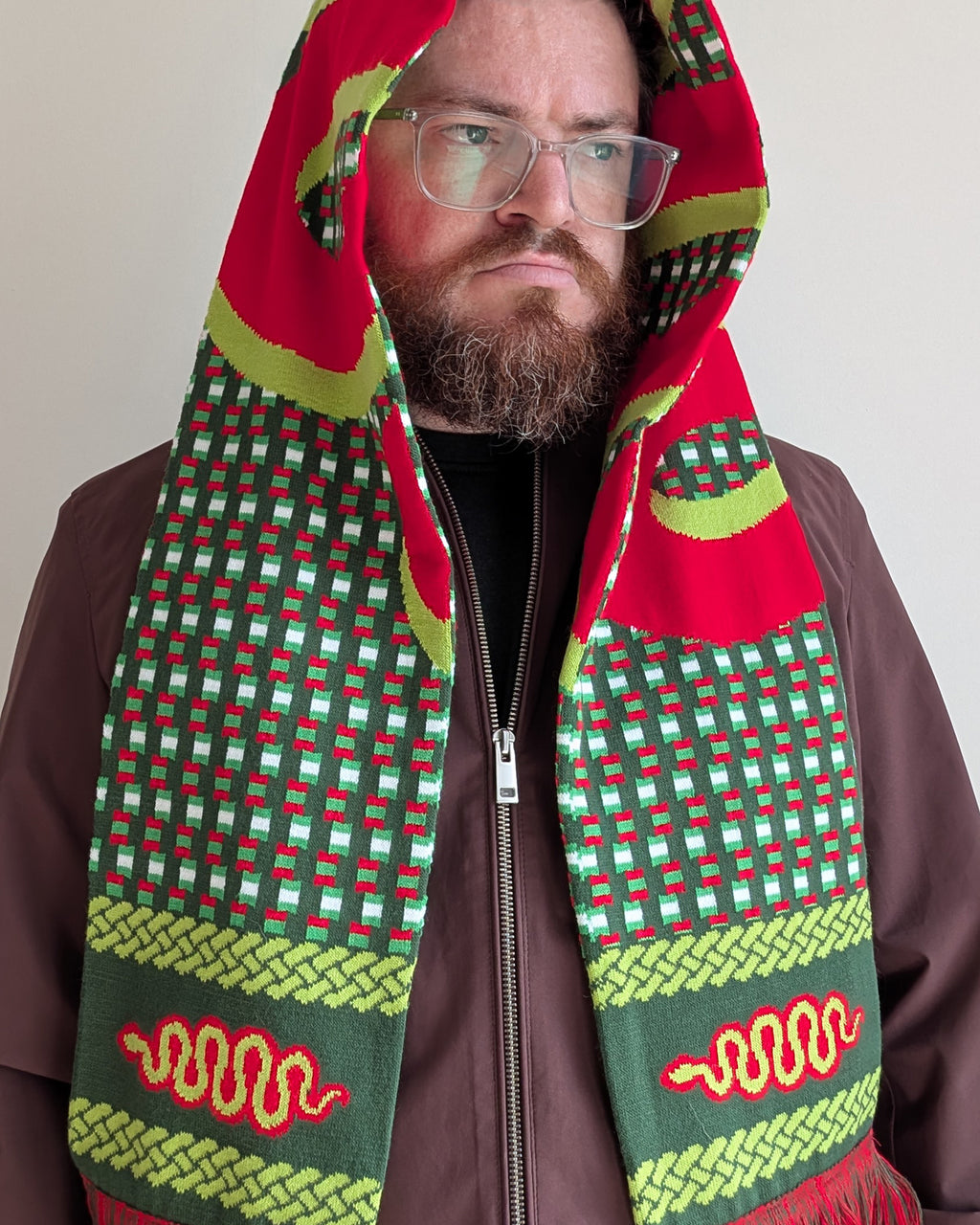 A man wearing a brown zip-up jacket and clear eyeglasses, with a red, green, and yellow patterned hooded scarf wrapped around his head and shoulders against a plain light background.