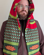 A man wearing a brown zip-up jacket and clear eyeglasses, with a red, green, and yellow patterned hooded scarf wrapped around his head and shoulders against a plain light background.