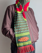 A person wearing a brown jacket and dark pants, shown from the chest down, wearing a vibrant green, red, and yellow patterned knit scarf with tassels.