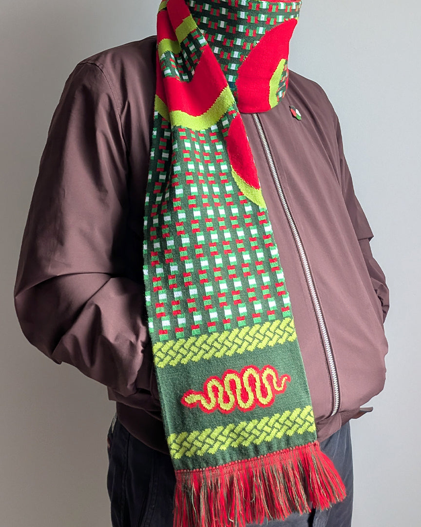 A person wearing a brown jacket and dark pants, shown from the chest down, wearing a vibrant green, red, and yellow patterned knit scarf with tassels.