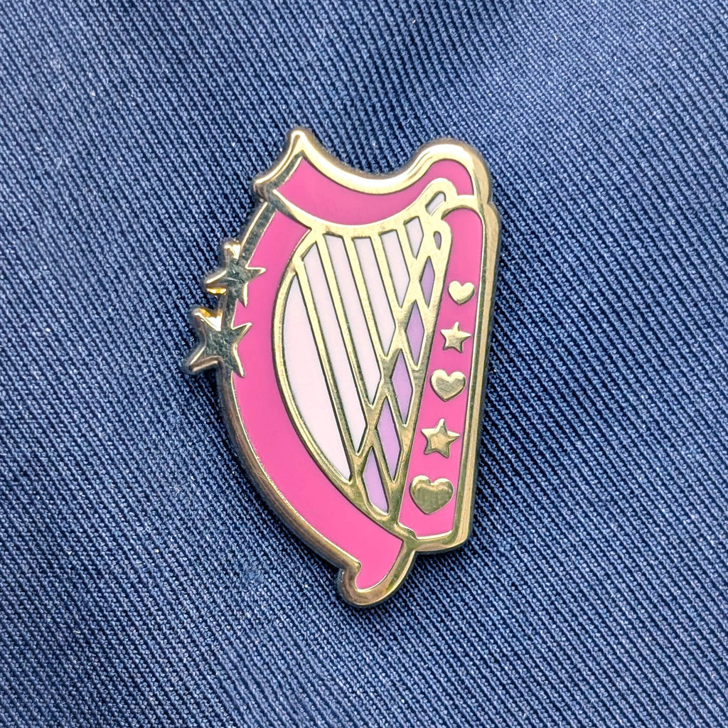 A pink and gold enamel pin shaped like a harp, featuring gold stars and hearts, displayed on a blue denim jacket background.