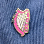 A pink and gold enamel pin shaped like a harp, featuring gold stars and hearts, displayed on a blue denim jacket background.