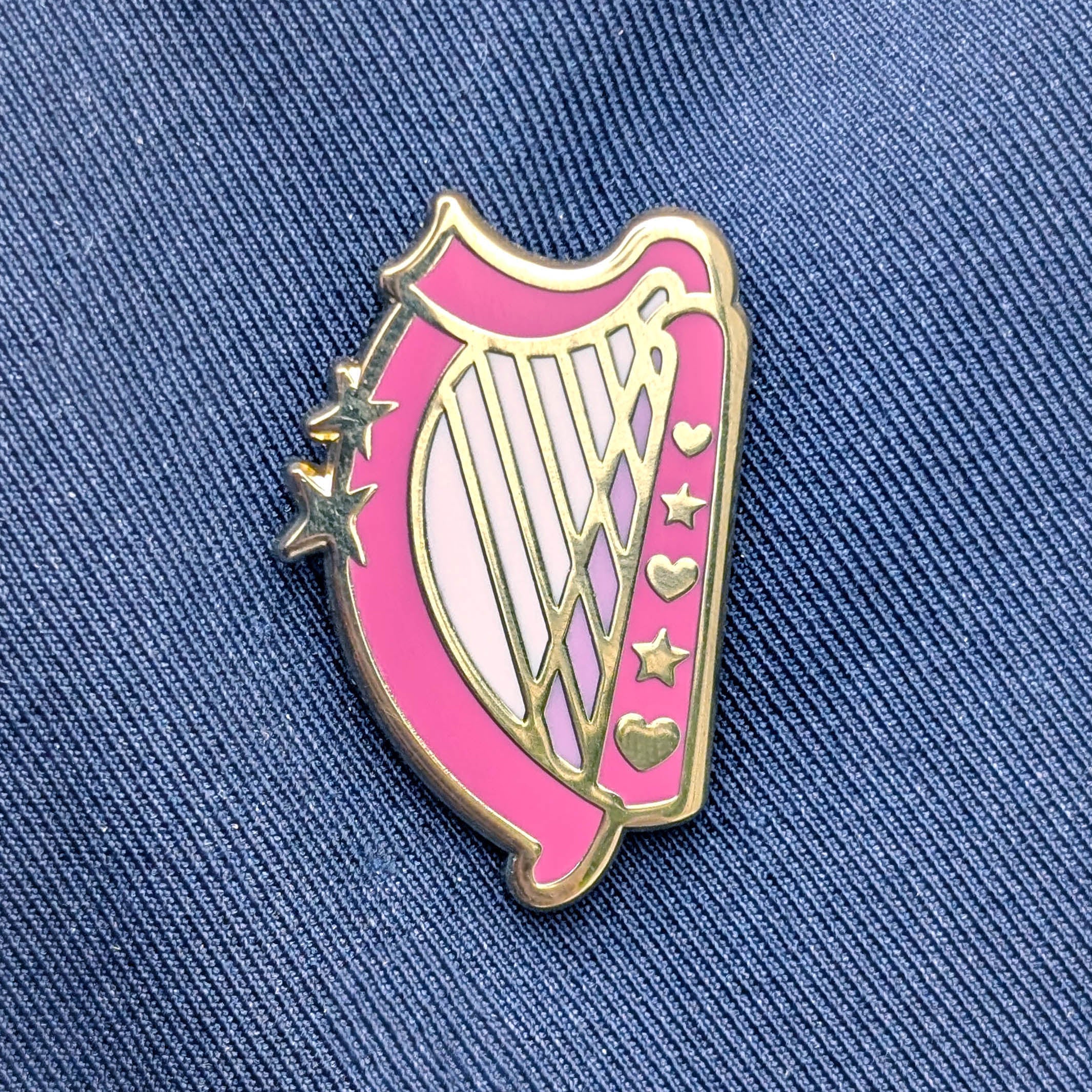 A pink and gold enamel pin shaped like a harp, featuring gold stars and hearts, displayed on a blue denim jacket background.