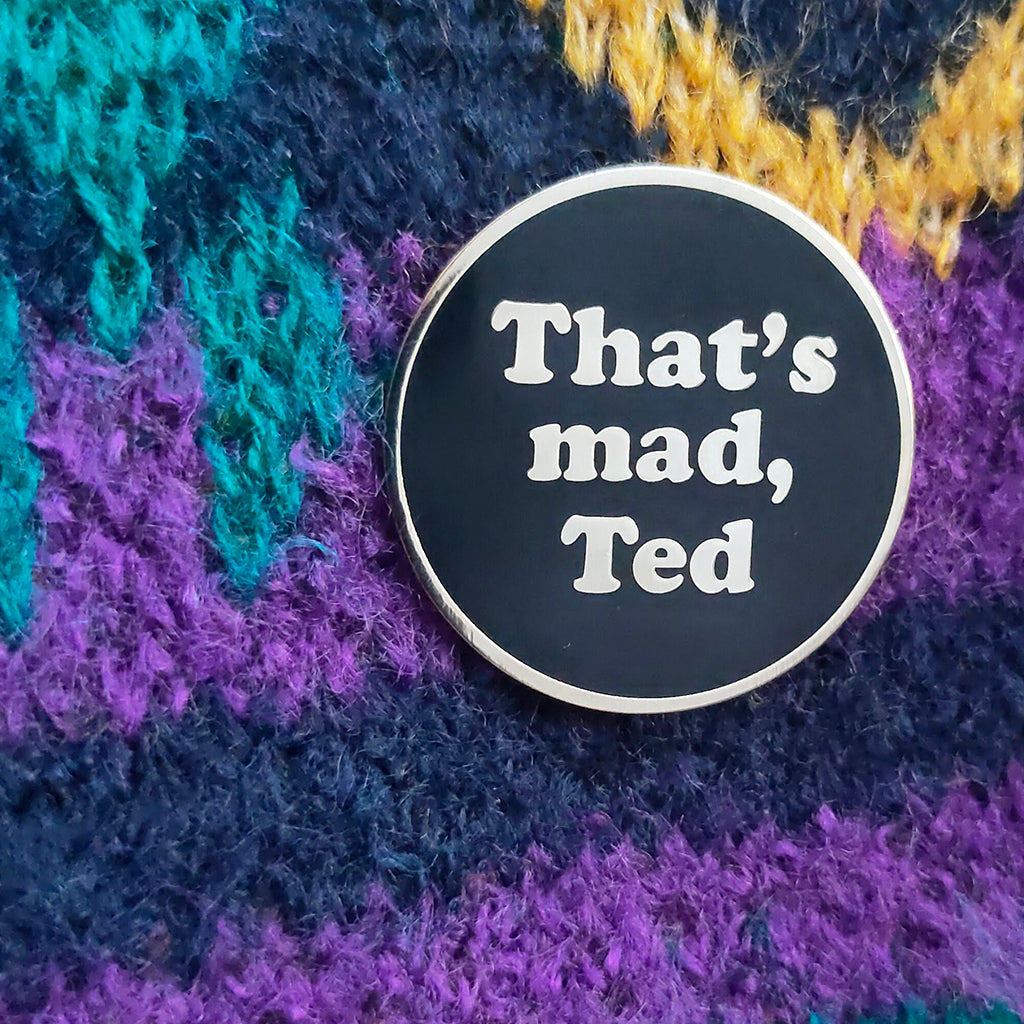 A round black enamel pin with a silver border and white text reading 'That's mad, Ted', placed on a textured multicolor knit fabric background.