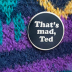 A round black enamel pin with a silver border and white text reading 'That's mad, Ted', placed on a textured multicolor knit fabric background.