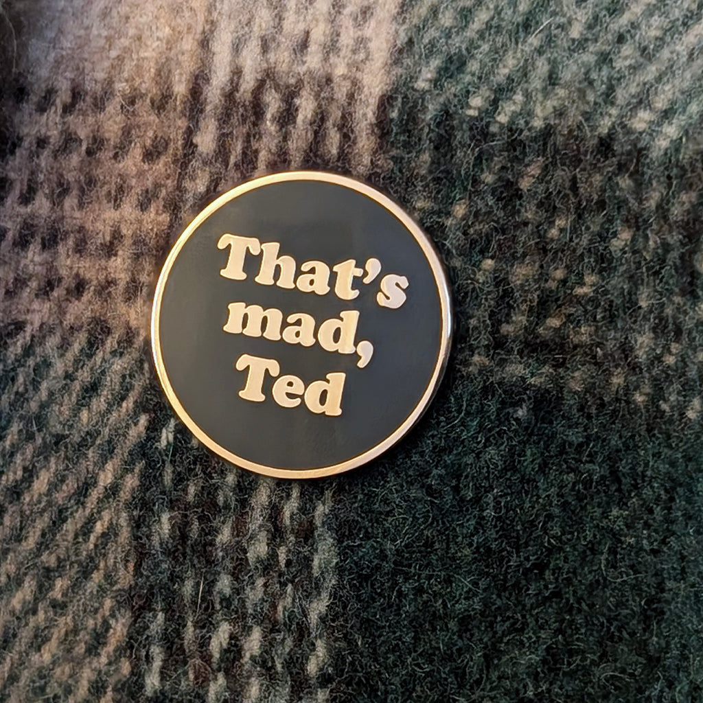 A round enamel pin with a black face and gold border, featuring the text 'That's mad, Ted' in gold lettering, placed on a dark green and gray textured knit fabric.