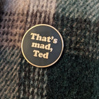 A round enamel pin with a black face and gold border, featuring the text 'That's mad, Ted' in gold lettering, placed on a dark green and gray textured knit fabric.